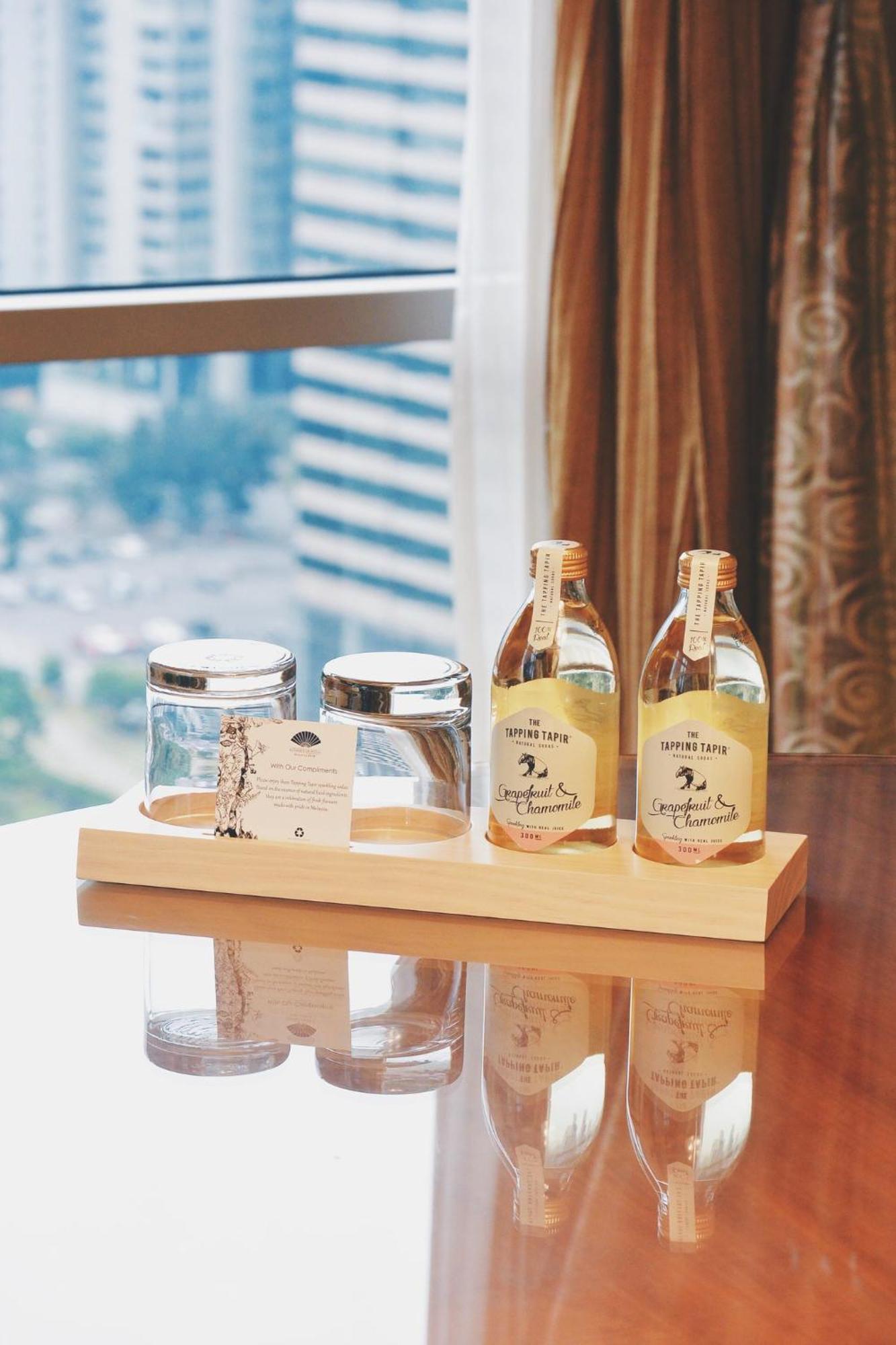 מלון Mandarin Oriental, קואלה לומפור מראה חיצוני תמונה The photo features a wooden tray displaying two glass bottles and two glass jars. The bottles contain a beverage, labeled "Sapphire Taps," with a note suggesting they are infused drinks. The jars are empty, with one having a metal lid. A small card i