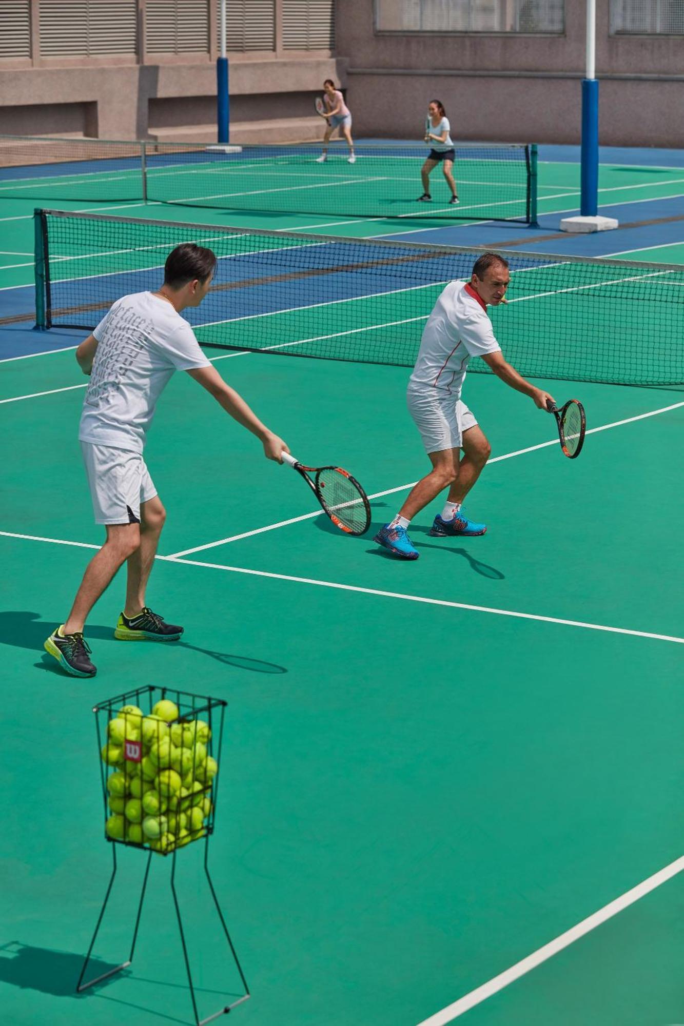 מלון Mandarin Oriental, קואלה לומפור מראה חיצוני תמונה The photo shows two men playing tennis on a green court. They are wearing matching white outfits and are actively hitting tennis balls with their rackets. In the foreground, there is a basket filled with yellow tennis balls. The court appears to have